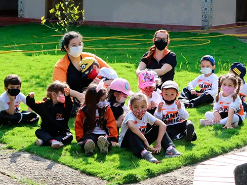 Um grupo de crianças e duas professoras sentadas em um gramado. Todos estão uniformizados e usando máscaras de proteção